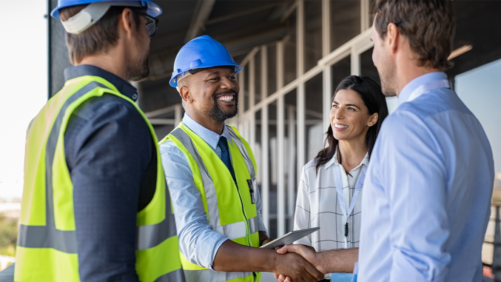 Man in construction hate shaking another man's hand with two onlookers.