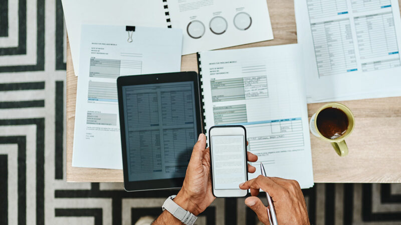 A person holds a cell phone over a desk of paperwork.